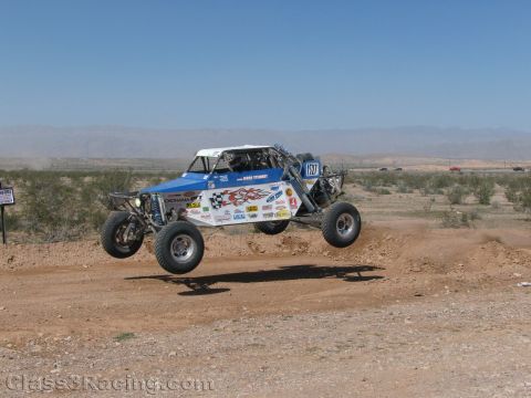 1990 Ford Bronco - blue-and-white off-road race buggy numbered 1271 catches air over a dirt berm in the desert.