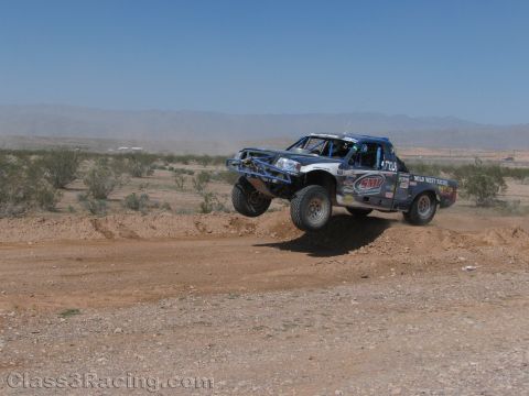 1990 Ford Bronco - white-and-blue off-road race truck with Wild West Racing livery leaps a dirt berm on a desert course.