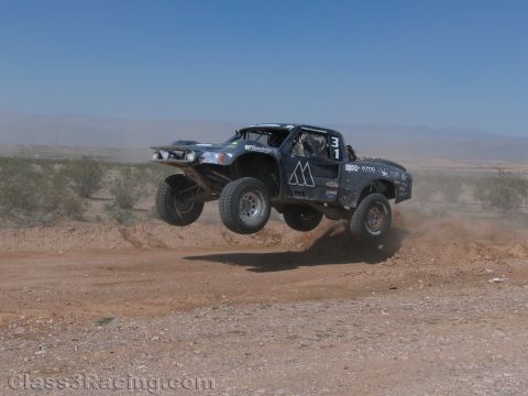 1990 Ford Bronco - dark off-road race truck numbered 31 kicks up dust as it jumps a dirt berm in the open desert.