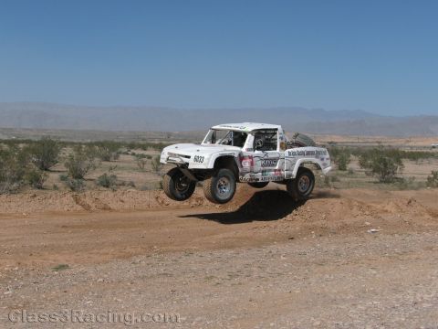 1990 Ford Bronco - white vintage Ford Bronco-style off-road race truck numbered 6603 catches air over a dirt berm in the desert.