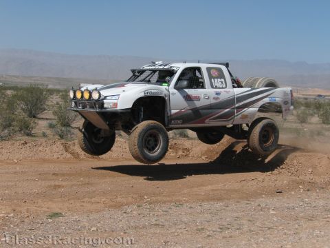 1990 Ford Bronco - white tube-chassis desert race truck numbered 1463 launches off a dirt berm in an arid mountain-backed landscape.