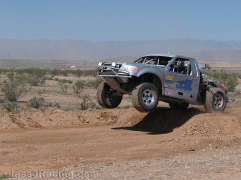 1990 Ford Bronco - silver tube-chassis off-road race truck jumps a dirt berm in the desert, dust trailing behind under a clear sky.