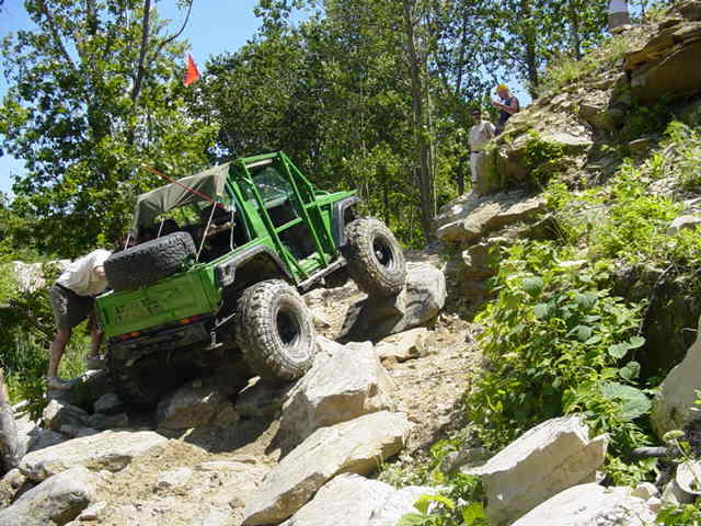 Green off-road vehicle with spare tire on back climbing a rocky hill. Person in tan shirt and shorts is near the rear