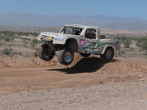 1990 Ford Bronco - white off-road race truck with a retro pickup body and KMC wheels catches air over a dirt mound in the desert.
