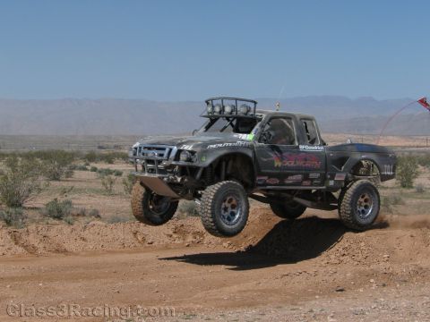 1990 Ford Bronco - dark Ford-badged compact race truck with roof basket lights jumps a dirt berm in a flat desert with distant mountains.