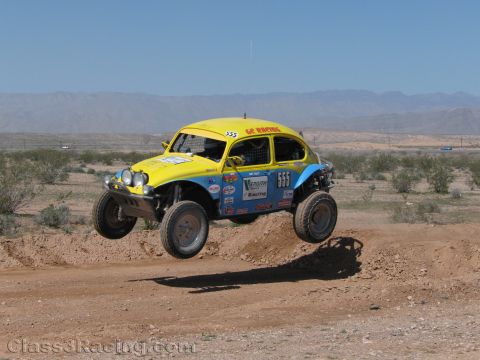1990 Ford Bronco - yellow Baja-style Volkswagen Beetle numbered 555 jumps a dirt berm, dust kicking up in the open desert.