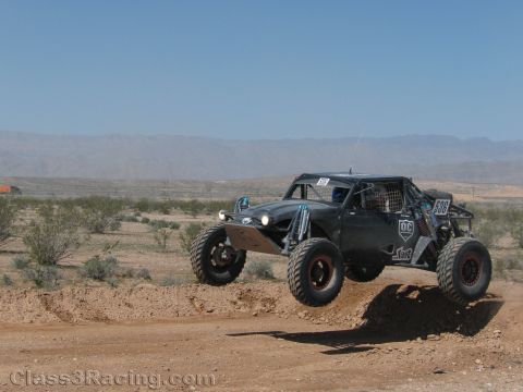 1990 Ford Bronco - black tube-chassis single-seat off-road buggy numbered 888 leaps a dirt jump under a clear blue desert sky.