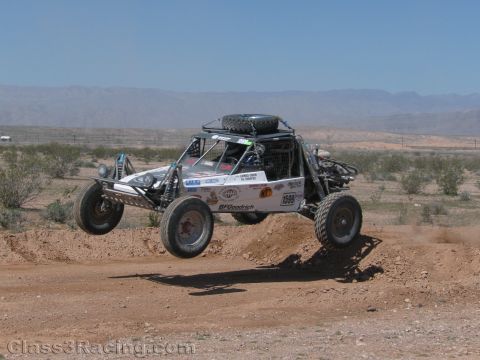 1990 Ford Bronco - open-cockpit tube-chassis off-road buggy numbered 1588, carrying a roof-mounted spare, catches air over a dirt mound.