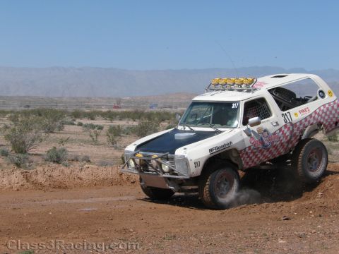 1990 Ford Bronco - white off-road race truck with red checkered graphics and No. 317 powers around a dirt corner, kicking up dust.