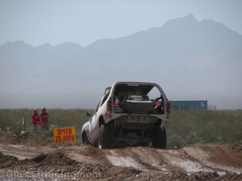 1990 Ford Bronco - Rear three-quarter view of a race truck numbered 317 driving a muddy desert course past a yellow 25 MPH speed sign.