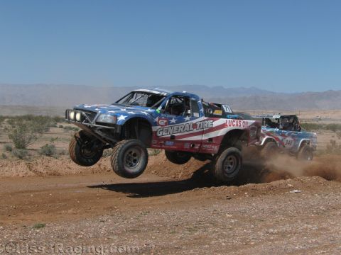 1990 Ford Bronco - American-flag-liveried General Tire race truck jumps a dirt berm in the desert as a second truck follows in dust.