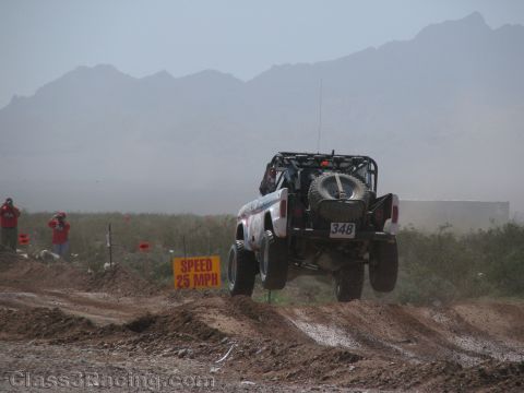 1990 Ford Bronco - Rear view of race truck No. 348 launching off a jump amid dusty haze, passing a yellow 25 MPH speed sign.