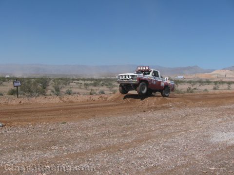 1990 Ford Bronco - white and red off-road race truck with roof lights crests a dirt berm on a wide desert course under a blue sky.