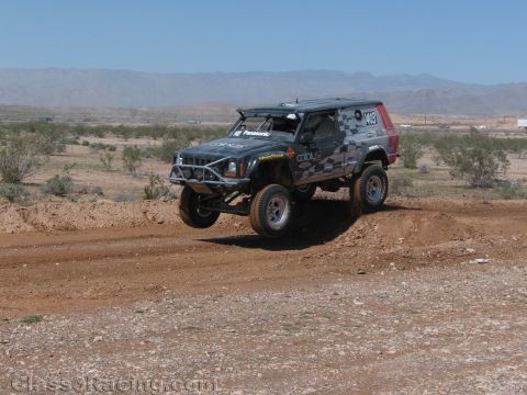 1990 Ford Bronco - gray camo-wrapped SUV-style off-road race truck climbs a dirt berm in the open desert with mountains behind.