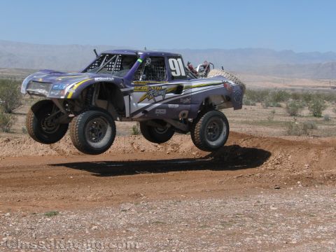 1990 Ford Bronco - purple and yellow off-road race truck numbered 91 catches air over a dirt jump in the desert under a clear sky.