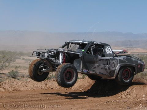 1990 Ford Bronco - dark camo-pattern No. 61 tube-chassis race truck leaps a dirt berm with long-travel suspension extended.