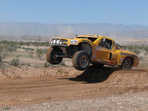 1990 Ford Bronco - yellow Toyo-Tires Green Bros race truck jumps a dirt berm in the desert, dust trailing under distant mountains.