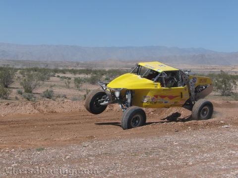 1990 Ford Bronco - yellow tube-chassis open-cockpit off-road buggy carves a dirt corner in the desert, kicking up dust.
