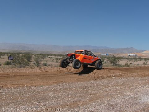 1990 Ford Bronco - orange tube-chassis off-road buggy catches air over a dirt berm on a wide flat desert course.