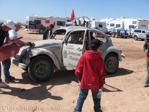 1990 Ford Bronco - white Johnson Racing Baja-style Volkswagen Beetle sits in a dusty pit area as crew and a boy in a red hoodie stand nearby.