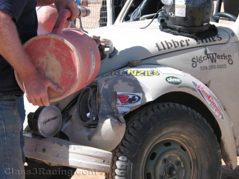 1990 Ford Bronco - person tips a red fuel jug into the fender area of a white Sign Werks-marked off-road race car during a pit stop.