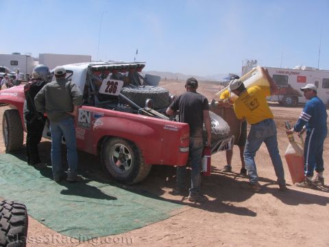 1990 Ford Bronco - pit crew services and refuels a red off-road race truck numbered 236 on a green mat in a dusty desert pit area.