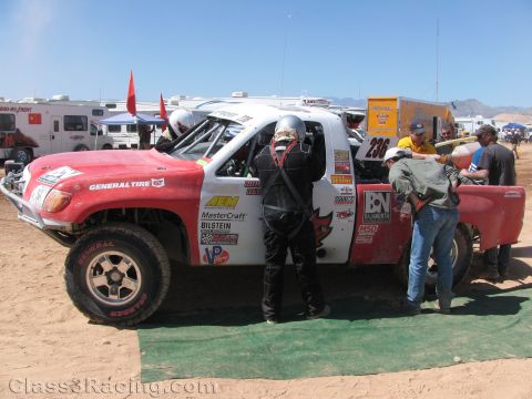1990 Ford Bronco - red and white General Tire race truck No. 236 sits in a desert pit as a helmeted driver and crew gather around it.