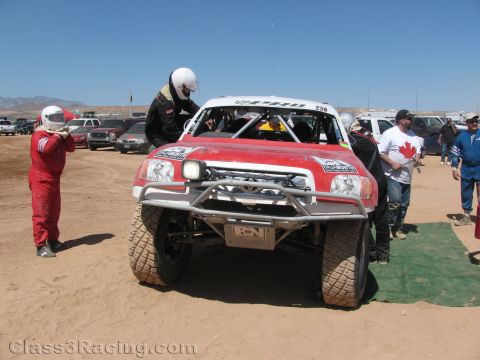 1990 Ford Bronco - Front view of a red and white off-road race truck numbered 238 surrounded by helmeted crew in a dusty desert pit.