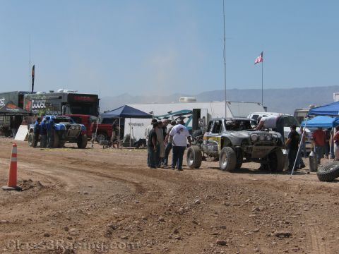 1990 Ford Bronco - busy desert race pit area with off-road trucks, canopy tents, RVs, spectators and an American flag flying overhead.