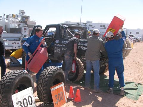 1990 Ford Bronco - pit crew in blue suits refuels and works on a black off-road race truck marked #300 at a desert race pit area.