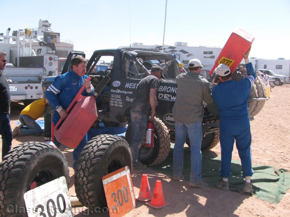 Crew in blue suits servicing black off-road race truck #300 in dirt pit