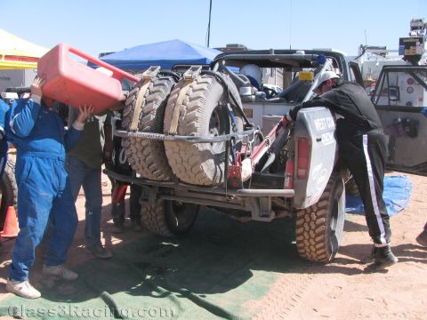 1990 Ford Bronco - crew member pours fuel from a red jug into the rear of an off-road race truck fitted with two mud-caked spare tires.