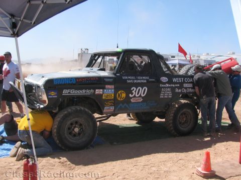 1990 Ford Bronco - black Bronco off-road race truck marked 300 sits under a canopy while crew members work beneath it in a dusty pit.