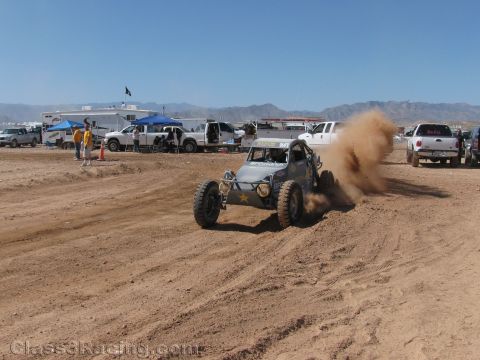 1990 Ford Bronco - gray single-seat off-road buggy throws up a plume of dust while racing on a dirt track past parked trucks.