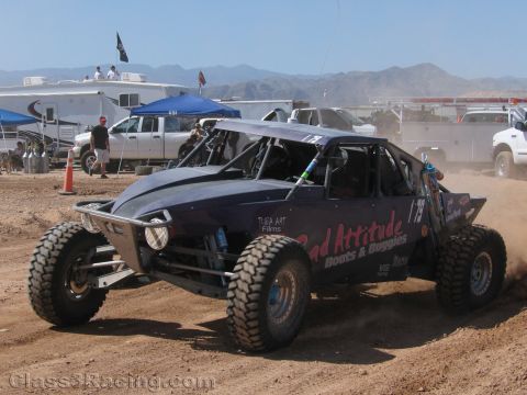 1990 Ford Bronco - dark single-seat off-road race buggy numbered 79 corners on a dirt track with RVs and mountains in the background.