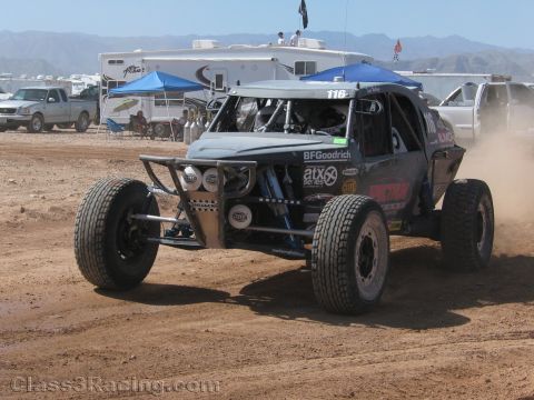 1990 Ford Bronco - gray off-road race buggy numbered 116 races across a dirt track in front of blue canopies and parked trucks.