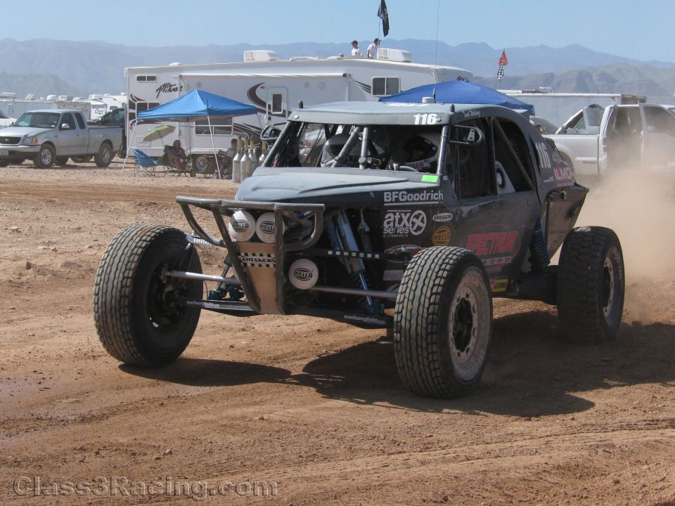 Gray off-road race buggy #116 on dirt track with blue canopies behind