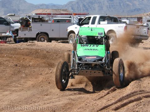 1990 Ford Bronco - green off-road race buggy numbered 1600 throws up dirt as it races past a white pickup truck on a dirt course.