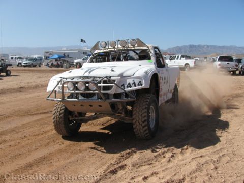 1990 Ford Bronco - white off-road race truck numbered 1414, topped with a roof light bar, kicks up dust on a desert dirt track.