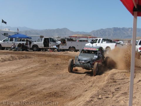 1990 Ford Bronco - dark off-road race buggy slides through a cloud of dust on a dirt track beside a row of pit trucks and a red canopy.
