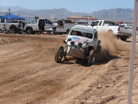 1990 Ford Bronco - light-colored off-road Baja-style Volkswagen Beetle buggy with American flag graphics races through dirt past parked trucks.