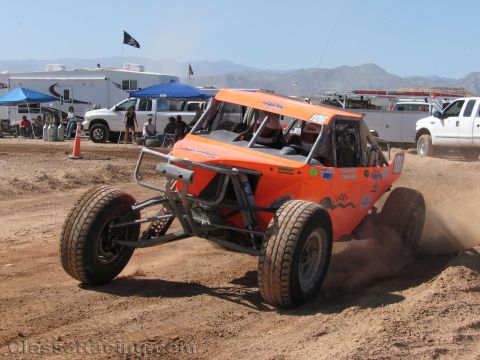 1990 Ford Bronco - orange single-seat off-road race buggy corners hard on a dirt track in front of parked trucks and blue canopies.