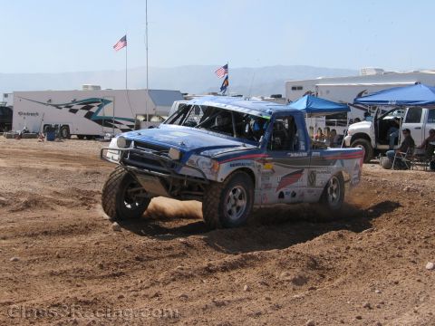 1990 Ford Bronco - blue and white off-road race truck turns on a dirt track with RVs, flags and tents lining the background.