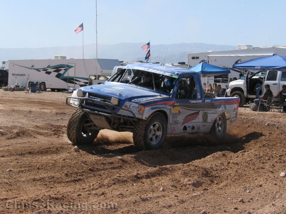 Blue and white off-road race truck turning on dirt with RVs behind