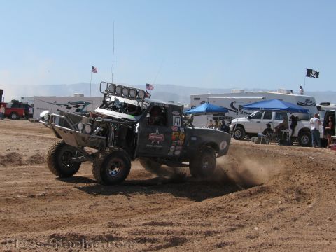 1990 Ford Bronco - silver off-road race truck numbered 57, fitted with a roof light bar, kicks up dust racing on a dirt course.
