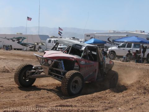 1990 Ford Bronco - red and silver off-road race buggy sprays a wave of dirt as it powers around a dirt track past parked vehicles.