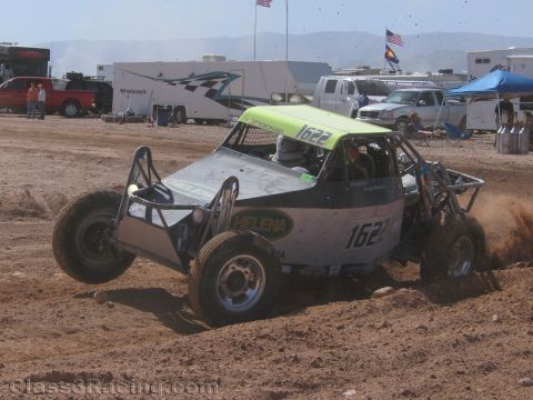 1990 Ford Bronco - gray off-road race buggy numbered 1622 with a green roof slides through dirt on a desert track near pit vehicles.