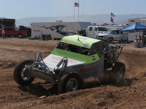 1990 Ford Bronco - green and white off-road race buggy corners on a dirt track in front of RVs, trucks and blue canopies.