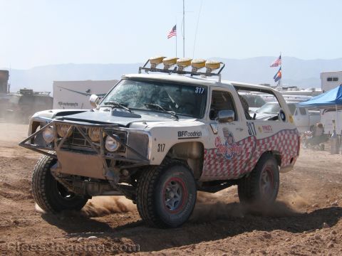 1990 Ford Bronco - white off-road race truck numbered 317 with red checkered graphics and a roof light bar kicks up dust on a dirt track.