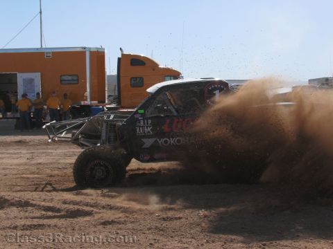 1990 Ford Bronco - black off-road race truck throws up a massive rooster tail of dirt beside an orange semi truck in a pit area.